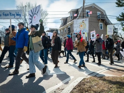 A group of people walk across a street during a protest, holding various signs including one reading "No Faux-King Way." Houses and power lines are visible in the background.