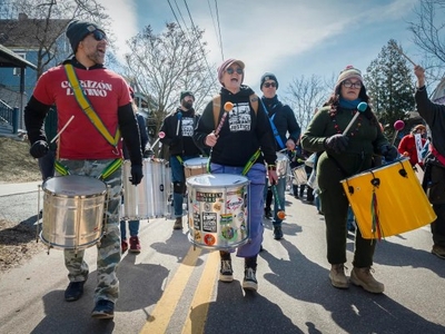 A group of people march down a street playing drums, wearing winter clothing and sunglasses, with trees and houses in the background.