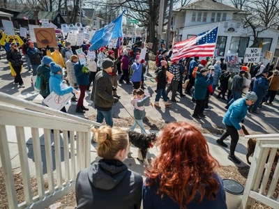 A crowd of people holding signs and flags gathers for a protest on a residential street, viewed from behind two seated individuals on a staircase.