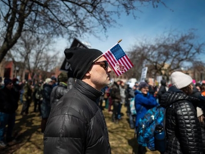 A man in a black beanie and sunglasses holds a small American flag at an outdoor gathering with other people on a grassy area.