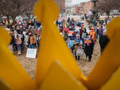 A crowd of people holding signs gathers outdoors on a cloudy day, partially obscured by a large yellow crown in the foreground.