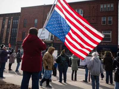 A group of people stand in a town square, some holding signs, while one person holds a large American flag. Brick buildings line the background.