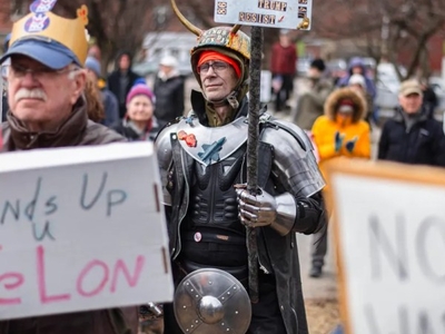 A group of protesters holds signs with messages like "NO WAR" and "Hands Up U Felon"; one person is dressed in medieval armor and a horned helmet.