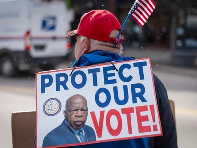 A person wearing a red cap with an American flag holds a sign reading "PROTECT OUR VOTE!" with a photo of John Lewis on their back. A USPS truck appears in the background.