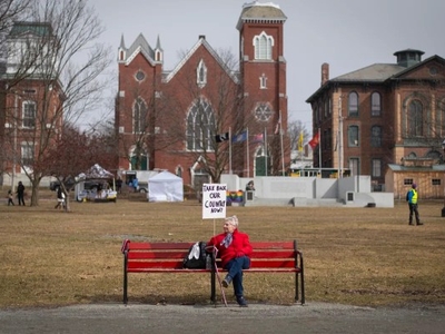 An older woman in a red coat sits alone on a red bench in a park, holding a sign that reads "TAKE BACK OUR COUNTRY NOW!" Buildings and tents are visible in the background.