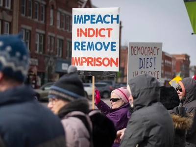 A group of people at a protest hold signs, including one that reads "IMPEACH INDICT REMOVE IMPRISON" in red and blue letters.