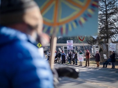 A group of people stands on a roadside holding protest signs, including one reading "NO KINGS," while a person in the foreground holds a sign with a peace symbol.