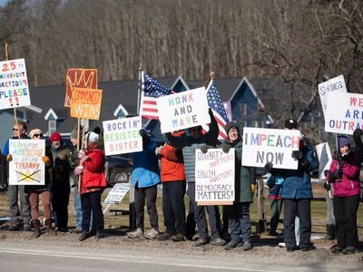 A group of people stand along a roadside holding protest signs with messages about impeachment, democracy, and political action on a sunny day.