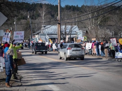 A group of people gather along a roadside holding various protest signs while cars drive by on a clear day.