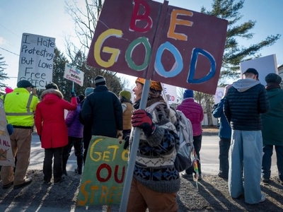 A group of people protest outdoors holding various signs, including one that reads "BE GOOD" and another that says "Get Off Our Streets.