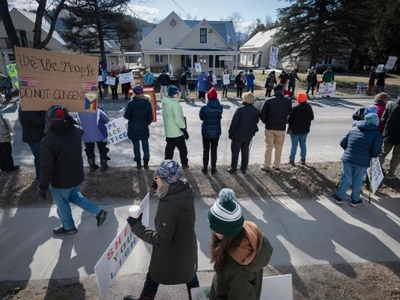 A group of people stand on a sidewalk and street holding signs during a daytime protest in a residential neighborhood.