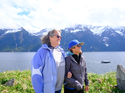 Sisters Sue York, executive director of the Eldred Rock Lighthouse Preservation Association (left) and Donna Lentz, who volunteered for the first lighthouse keeper shift, watch the flag rising during the opening ceremony of the lighthouse on May 30, 2026. (Photo by Corinne Smith/Alaska Beacon)
