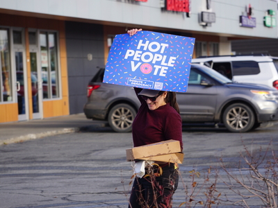 A No Kings participant holds a sign and a box of donuts at a gathering site in Midtown Anchorage on Oct. 19, 2026. (Photo by Yereth Rosen/Alaska Beacon)