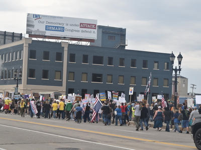 Under a League of Women Voters sign, a long line of people march down a sidewalk across a bridge.