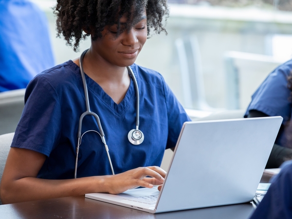 In class, a diligent female nursing student in scrubs engages in the discussion and takes notes on her laptop simultaneously.