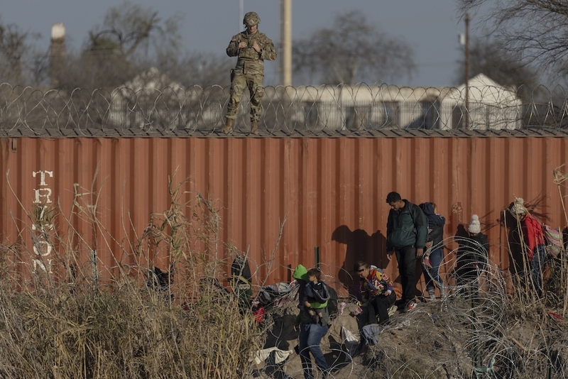 A National Guard soldier stands on top of shipping containers and watches as migrants traverse concertina wire at the bank of the Rio Grande in Shelby Park at Eagle Pass, Texas, on Jan. 19, 2024. (Photo by Eddie Gaspar / The Texas Tribune)