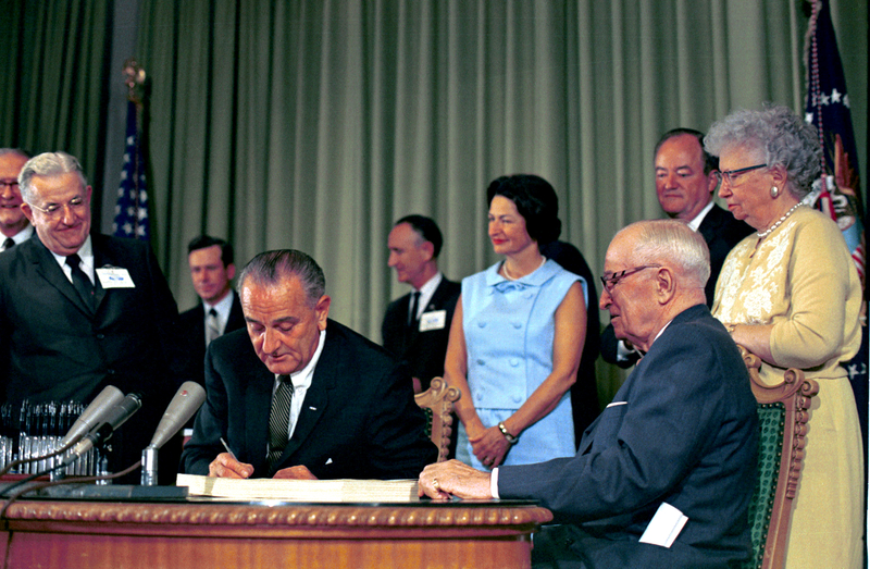 President Lyndon B. Johnson in 1965 signs the bill into law that established Medicare and Medicaid. Bess Truman as well as former President Harry S. Truman and Lady Bird Johnson and others look on. (LBJ Presidential Library photo)