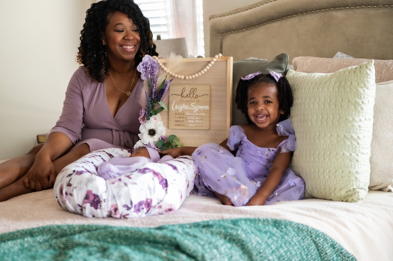 A mom and daughter sit on a bed with a new baby sibling in this posed photo.