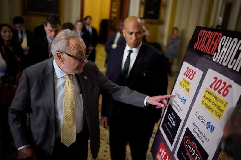 Senate Minority Leader Chuck Schumer, D-N.Y., left, accompanied by Sen. Cory Booker, D-N.Y., points to a poster depicting rising medical costs if Congress allows the Affordable Care Act tax credits to expire in December as he speaks to reporters following a Democratic policy luncheon at the U.S. Capitol on Oct. 15, 2025 in Washington, D.C. (Photo by Andrew Harnik/Getty Images)