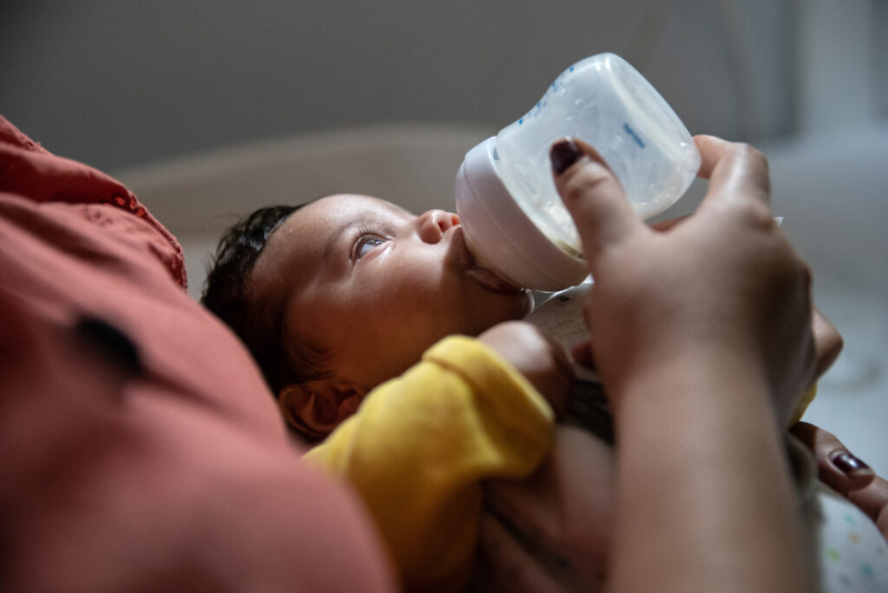 A baby drinks from a bottle while looking away.