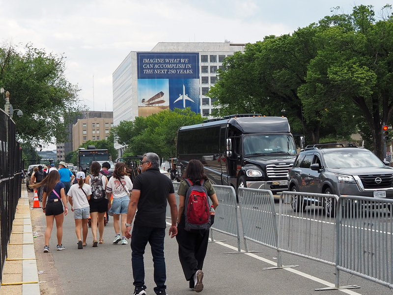 Tourists walk along the barricades surrounding the Freedom 250 construction site on 7th Street NW on Monday, June 22, 2026. A banner in the distance promoting Freedom 250 hangs on the Federal Aviation Administration headquarters. (Photo by Ashley Murray/States Newsroom)