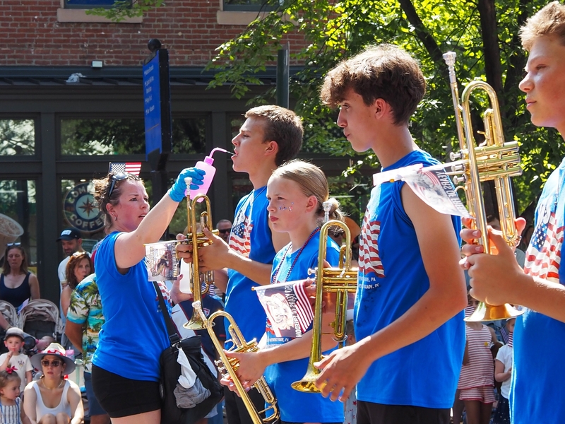 A chaperone from Nebraska squirted water into the mouth of a Grand Island Senior High School marching band member at the Capitol Hill Fourth of July Parade on Saturday, July 4, 2026 in Washington, D.C. The band marched in the smaller parade after the National Independence Day Parade was canceled due to the extreme heat. (Photo by Ashley Murray/States Newsroom)