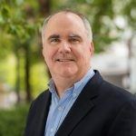 A photo of Tim Whitehouse, a white man who is balding, wearing a light blue dress shirt and a navy blazer.