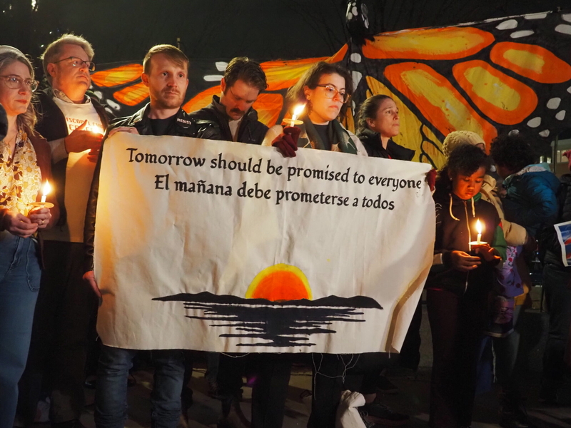 Several people hold candles as well as a banner reading “Tomorrow should be promised to everyone” in English and Spanish.
