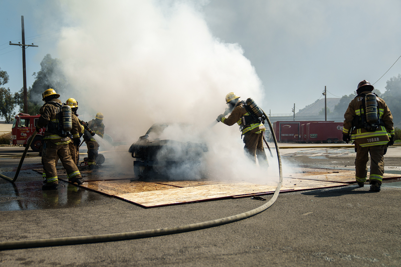 Firefighter trainees work to extinguish all flames on a burning car as smoke billows out of the vehicle.