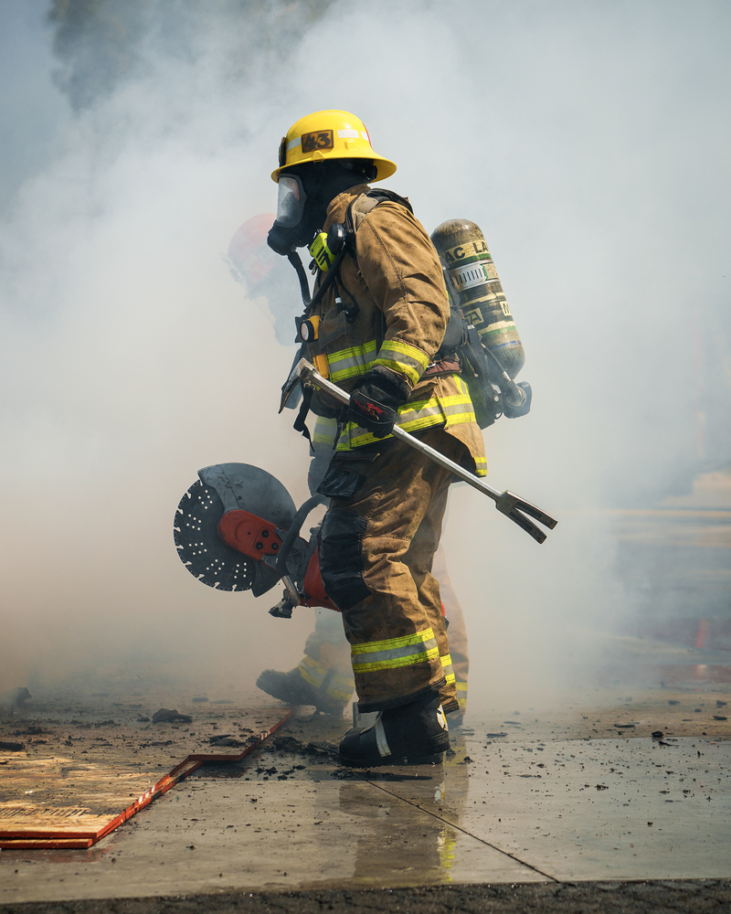 A firefighter trainee carries a saw during a car fire training session while surrounded by smoke.