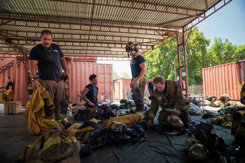 Several training captains remove their firefighting equipment and rest under a shaded canopy.