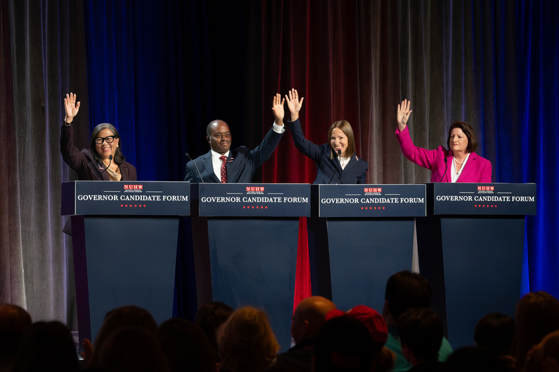 Four people stand on a stage behind individual podiums, each of them holding one hand up in the air in front of an audience at a governor candidate forum. Blue, red and gray lights illuminate the stage curtains the background.