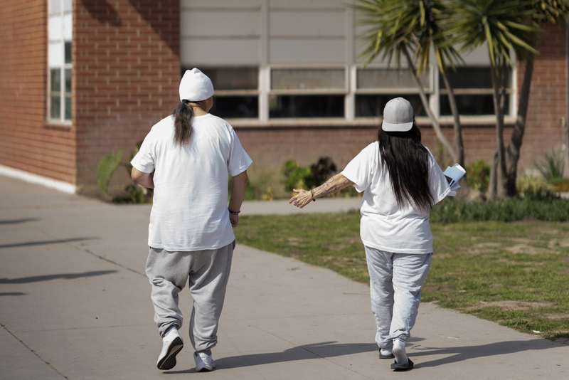 Two inmates dressed in gray sweatpants and white t-shirts walk towards a building on the grounds of a women's prison.