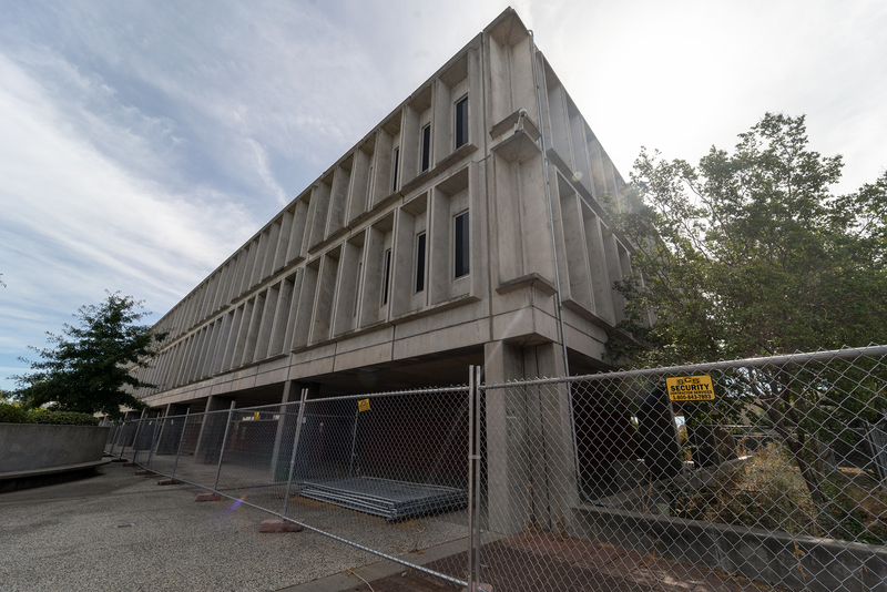 A concrete, modernist-style building with vertical windows stands behind a chain-link security fence. A &amp;#039;SEC Security&amp;#039; sign is visible on the fence, and construction materials are stacked behind it. The building is two stories tall with deep-set window recesses, and the area surrounding it closed off. Trees and cloudy skies are in the background, with sunlight shining through the branches.
