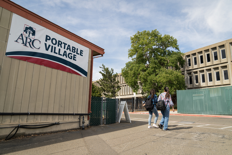 A portable building with a sign that reads &amp;#039;ARC Portable Village&amp;#039; is in the foreground on the left side of the image. Two individuals, wearing backpacks, walk towards a gated area with a modernist concrete building in the background. Trees surround the area, and the sky is clear with some clouds.