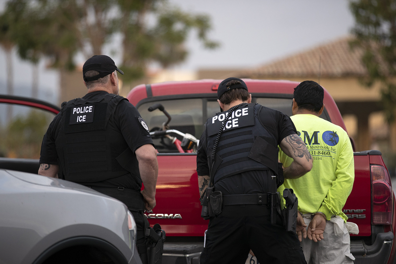 An officer searches a man in a yellow shirt handcuffed and leaning against a red truck, as another officer watches them.