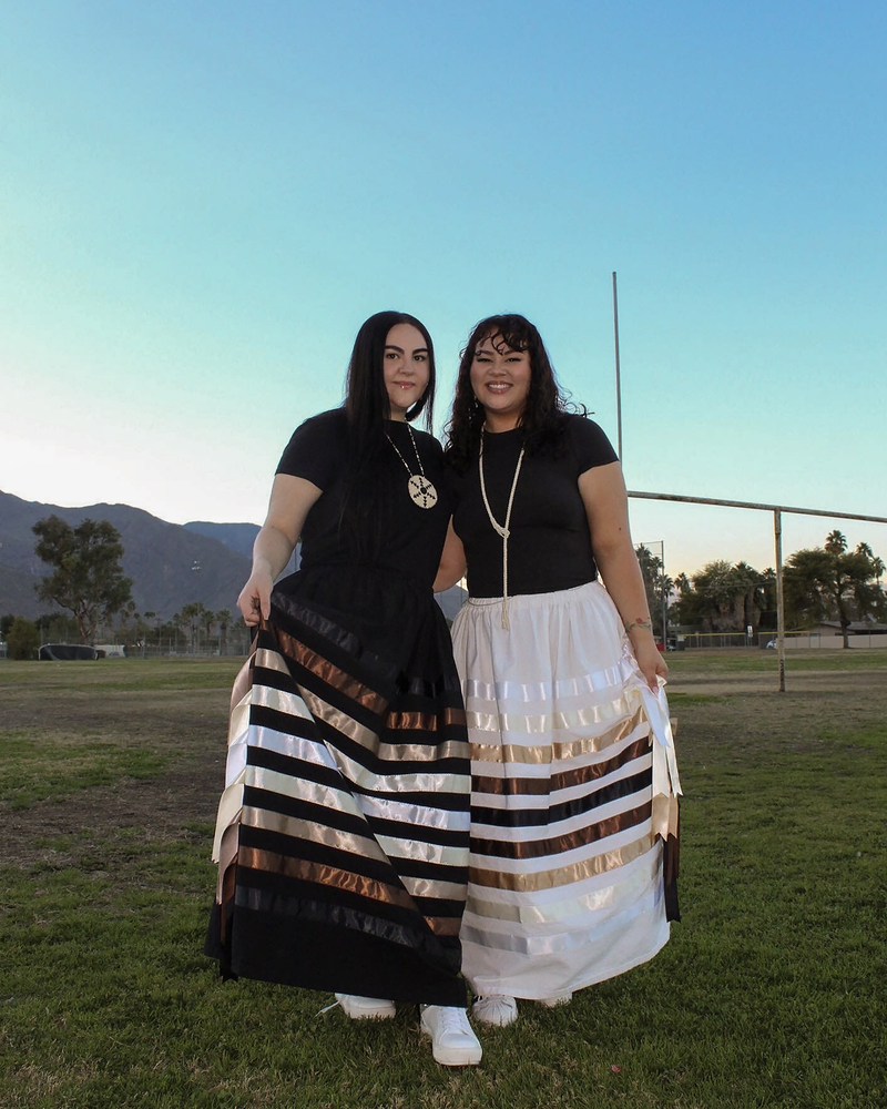 Two individuals stand together outdoors on a grassy field during dusk, with mountains and trees in the background. Both are wearing matching black tops and long ribbon skirts featuring metallic stripes, accessorized with necklaces. They are smiling and holding the edges of their skirts, showcasing the designs.