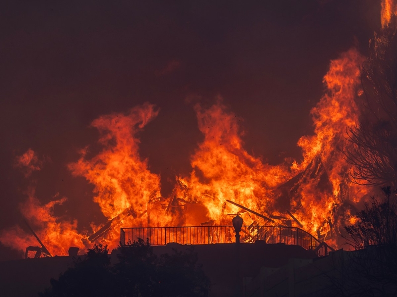 A raging wildfire consumes a structure, with massive flames engulfing the scene. Bright orange and yellow fire contrasts against the dark sky, creating an intense and dramatic visual. A fence and some vegetation are silhouetted in the foreground, emphasizing the destructive power of the blaze.