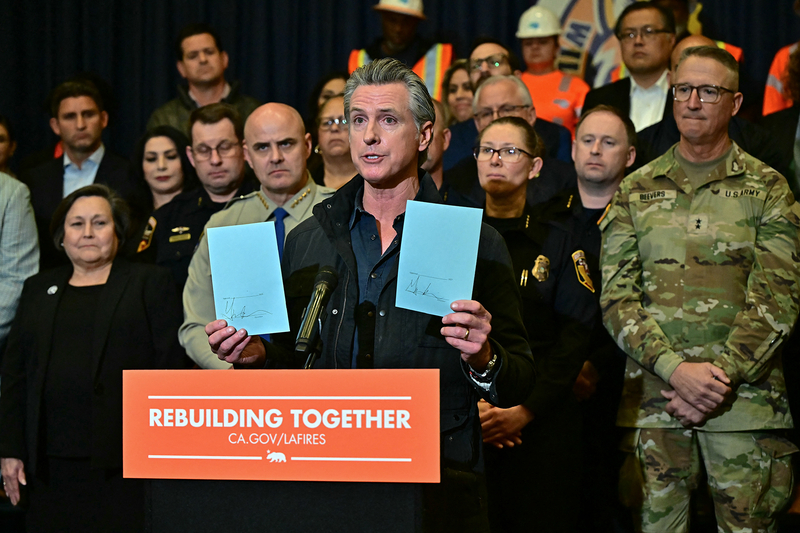 A person, wearing a black jacket and navy blue shirt, stands behind a lectern holding two pieces of paper on either hand. A group of lawmakers and public officials stand behind.