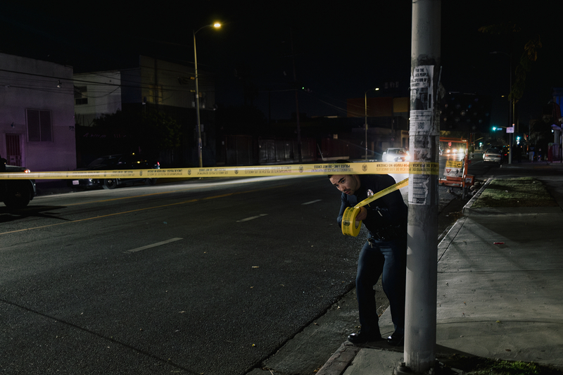 A police officer crouches down as they wrap yellow police tape around a light post in a street during an active crime scene on a night in Los Angeles.