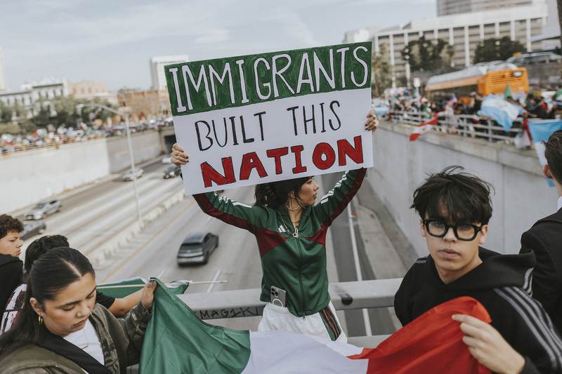 A person in a green tracksuit holding up a sign saying, “IMMIGRANTS UILT THIS NATION,” while next to a guard railing over a highway in downtown Los Angeles. In the background, cars can be seen driving on the road while crowds of protesters hold up signs and flags on both sides of the highway.