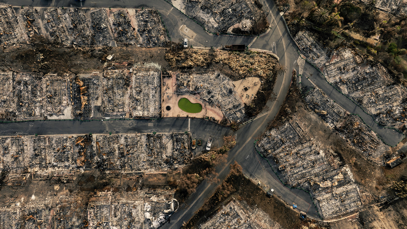 An aerial view of a residential area shows widespread destruction, with rows of homes reduced to charred rubble. Streets wind through the remains, and a small, murky green pool stands out amid the devastation. Scorched trees and debris surround the site, indicating the aftermath of a large fire.