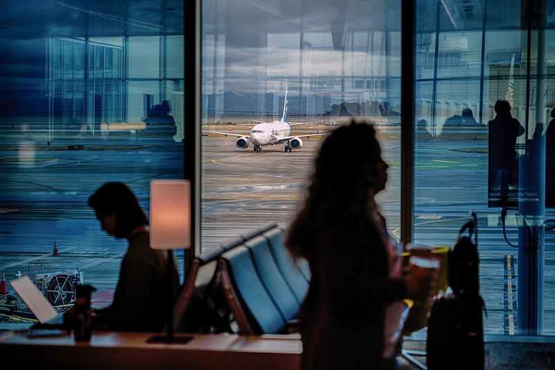 A view of a plane on the tarmac of an airport through the windows of a terminal. In the foreground of the photo is two silhouettes of people in the airport, one sitting down working at their laptop while the other walks by with their luggage. In the reflection of the glass, more people can be seen waiting in the airport terminal.