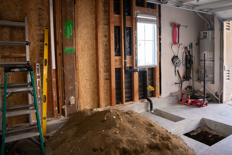 A medium-sized pile of dirt sits in the middle of a garage under construction. Next to the pile of dirt are two cemenet holes with plumbing pipes.