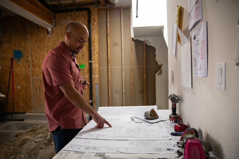 A person wearing a dark pink collared shirt and blue jeans uses their right index finger to point to floor plans laid on top of a white table inside a garage under construction.
