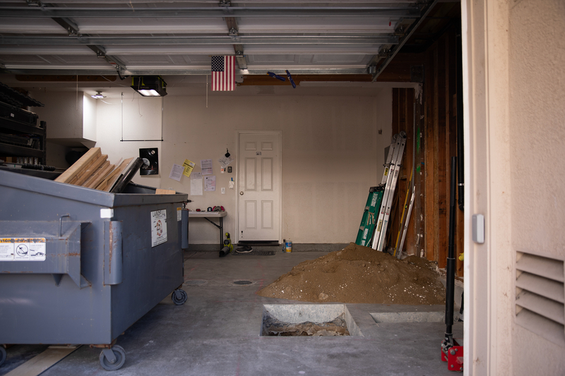 The inside of a grage under construction that includes a pile of dirt, a large grey garbage container and ladders.