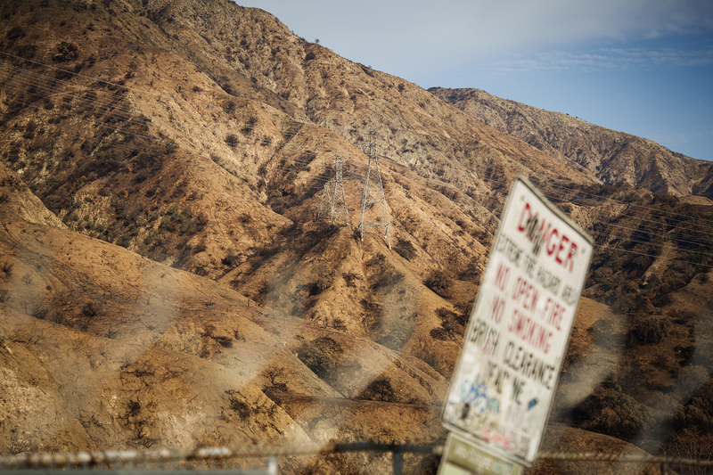 A landscape of scorched hills shows remnants of a wildfire, with dry, barren slopes dotted with blackened vegetation. Power lines and transmission towers stretch across the terrain. In the foreground, a blurred warning sign with red lettering reads "DANGER" and cautions against open fires and smoking. The hazy fence adds a sense of distance, emphasizing the vast, rugged expanse of the burned landscape under a clear sky.