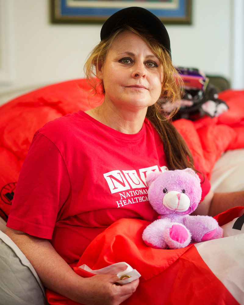 A person wearing a black hat and a red t-shirt holds pink teddy bear on their lap as they look directly into the camera.