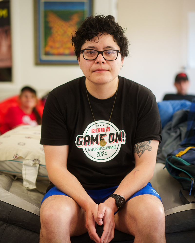 A person with short and curly black hair and wearing glasses with a black t-shirt sits at the edge of a mattress while looking into the camera.