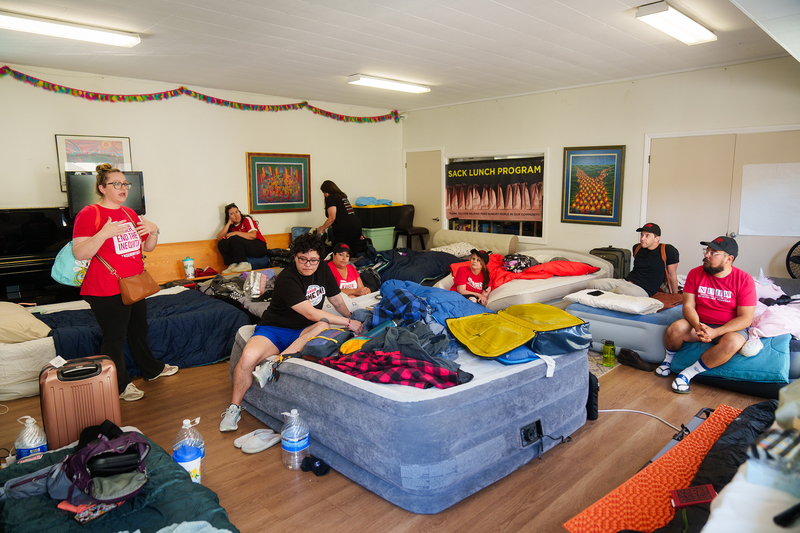 People wearing red or black t-shirts gather in a room with air mattresses and other personal belongings while they participate in a hunger strike.